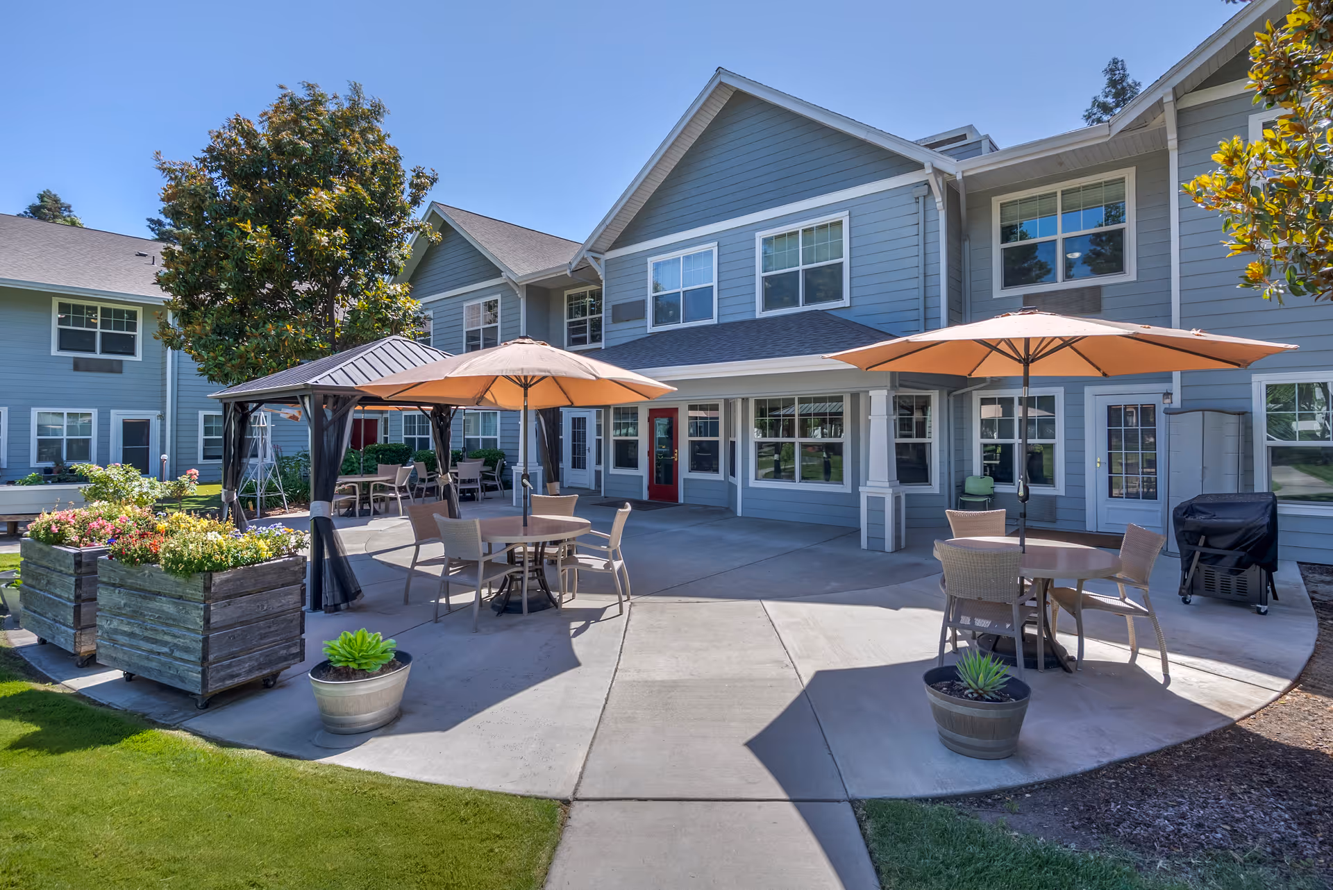 Sunny courtyard of a senior living facility with patio tables, umbrellas, planters, and the building façade in the background.