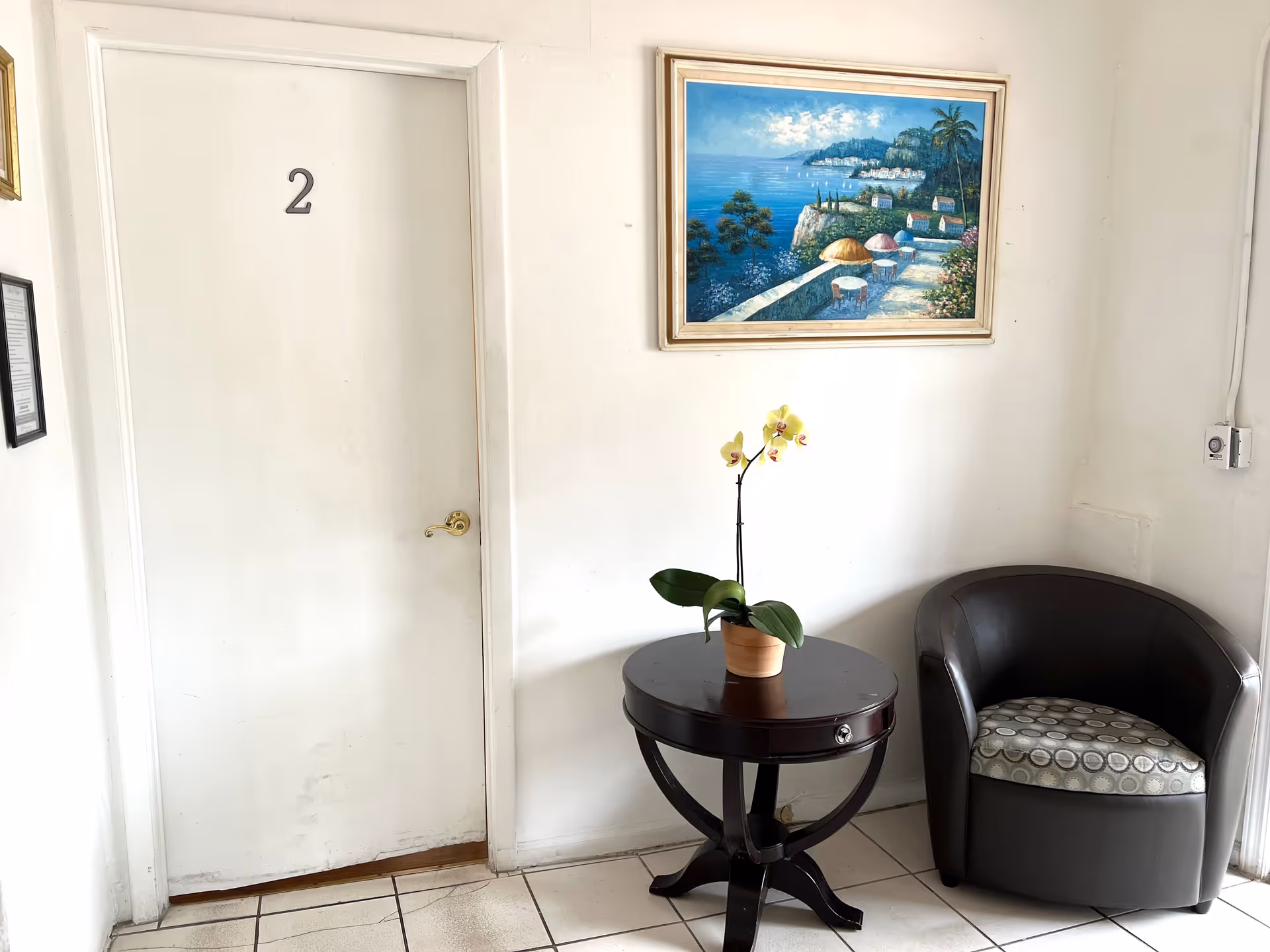 A small waiting area with a white door labeled number 2, a round dark wooden table with a potted orchid plant on it, a black cushioned armchair with a patterned seat cushion, and a framed painting of a coastal scene hanging on the white wall.