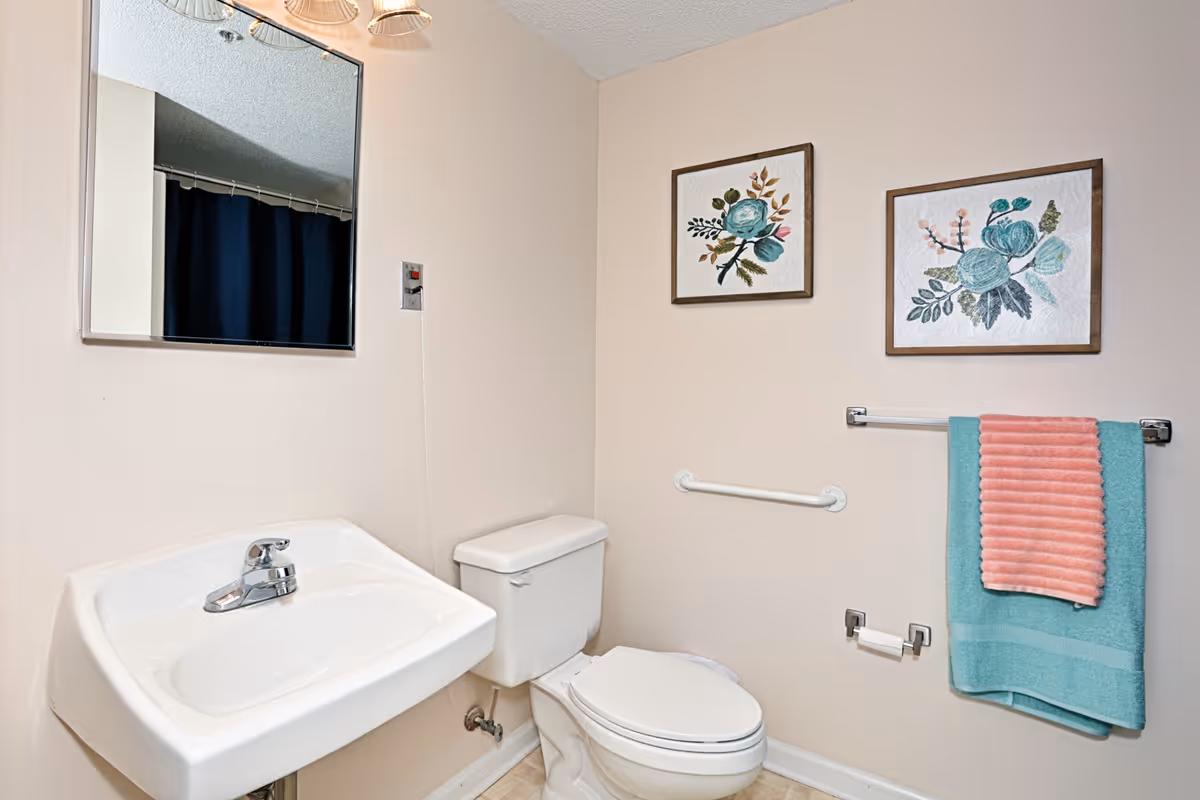 Small bathroom with a wall-mounted sink, toilet, mirror, floral wall art, and teal and coral towels on a rack.