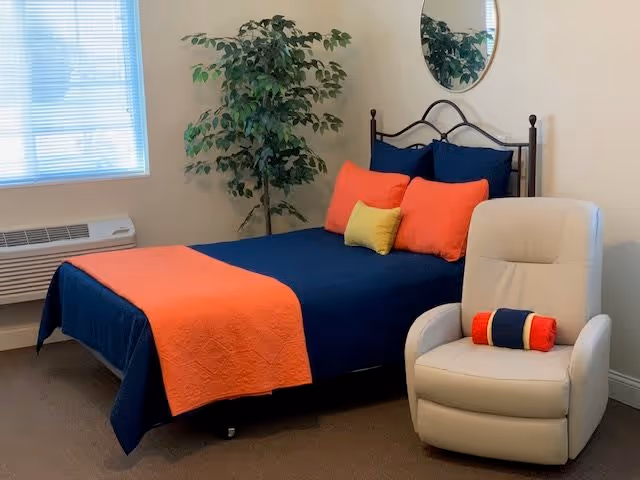 A bedroom with a bed dressed in navy blue bedding and orange and yellow accent pillows and blanket. There is a beige recliner chair with a small rolled towel on it, a large green potted plant in the corner, a window with blinds, and a round mirror on the wall.