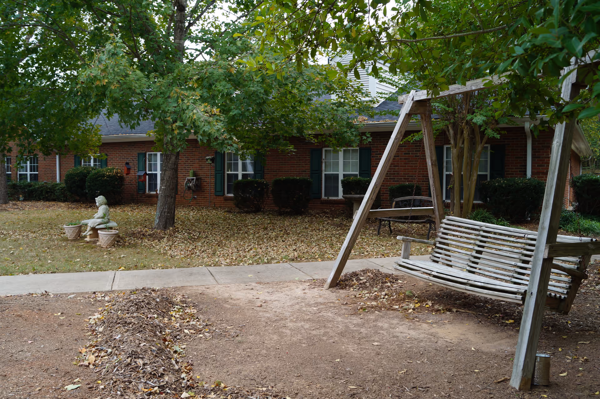 Outdoor area of a senior living facility with a wooden swing bench under a tree, a concrete pathway, a statue of a child sitting on a bench, and a brick building with green shutters in the background surrounded by bushes and fallen leaves.