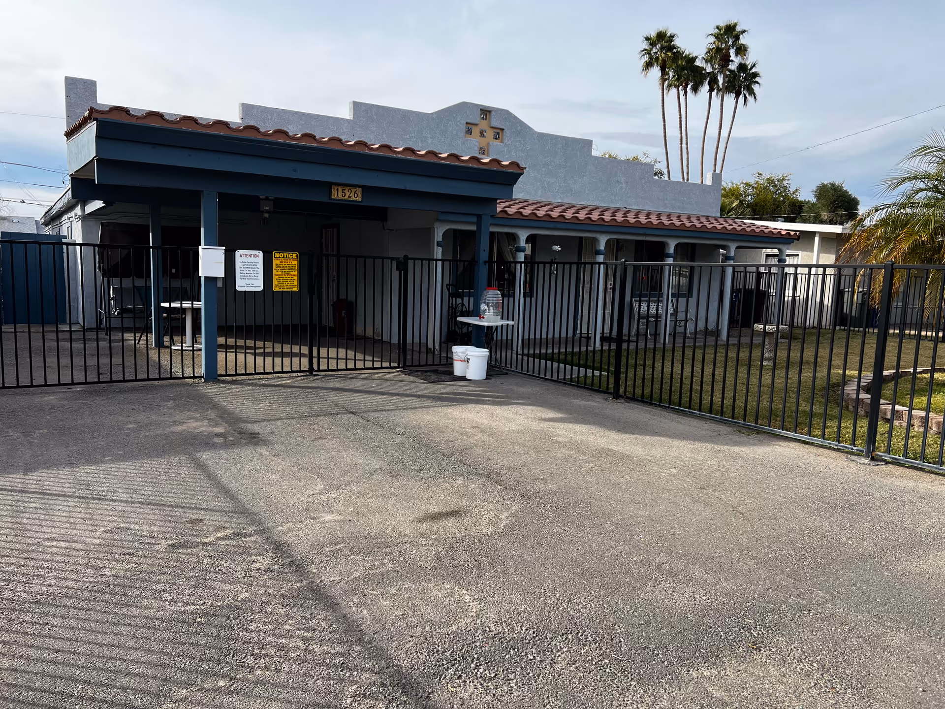 Exterior view of a single-story building with a gated entrance and a covered porch area. The building has a tiled roof and a decorative cross on the facade. There are palm trees and some greenery visible in the background.