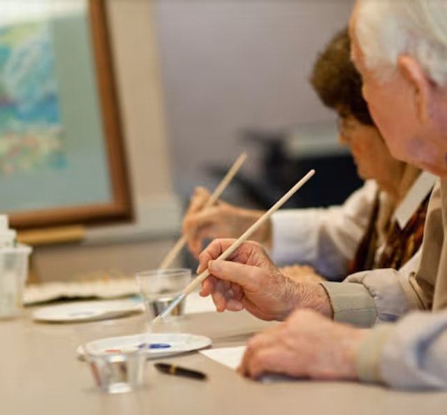 Two elderly individuals seated at a table, each holding a paintbrush and engaging in a painting activity with paint palettes and water cups in front of them.