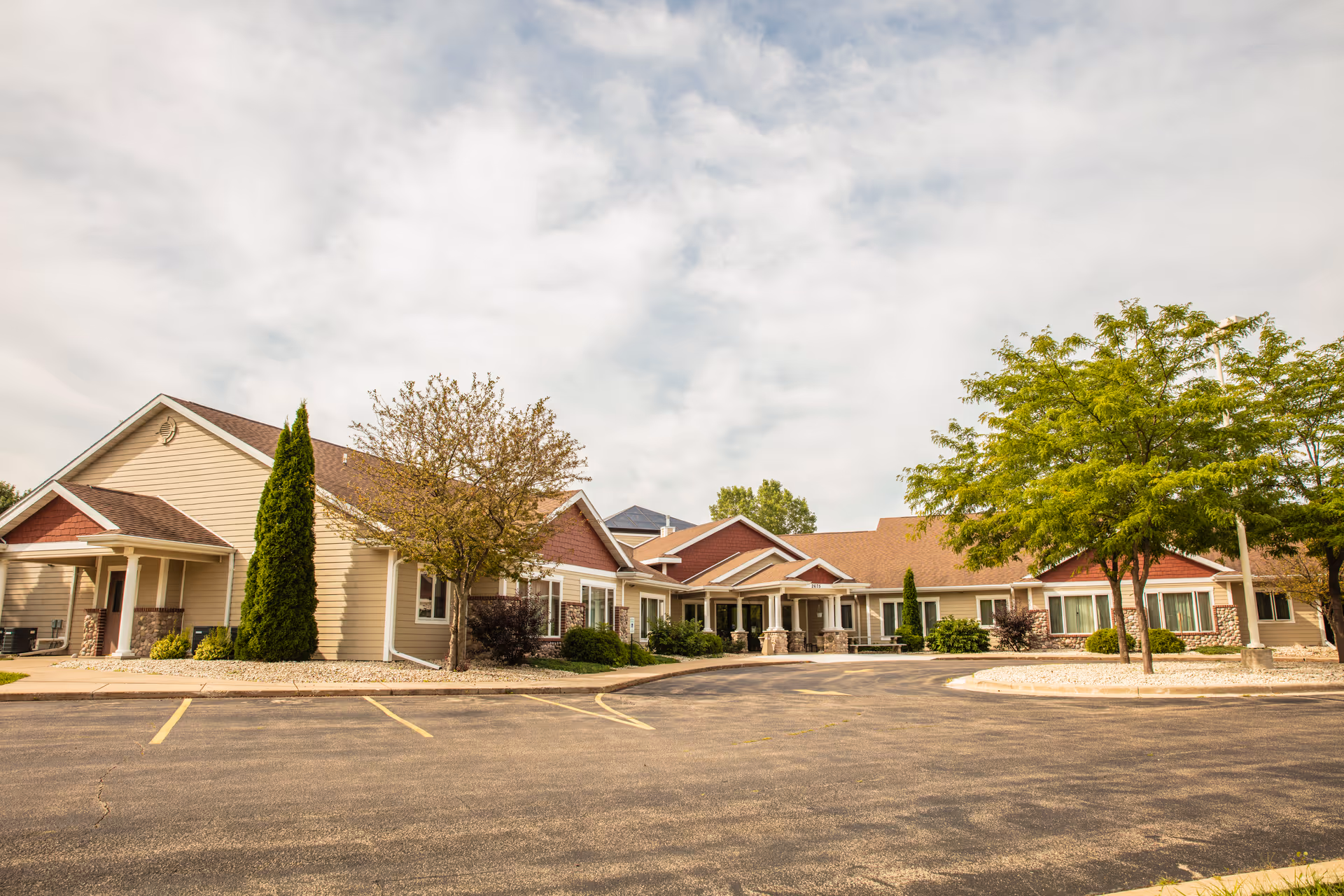 Exterior view of Oakwood Manor, a single-story senior living facility with beige siding, red accents, and a brown roof. The building is surrounded by trees and landscaping, with a paved driveway and parking area in front under a partly cloudy sky.