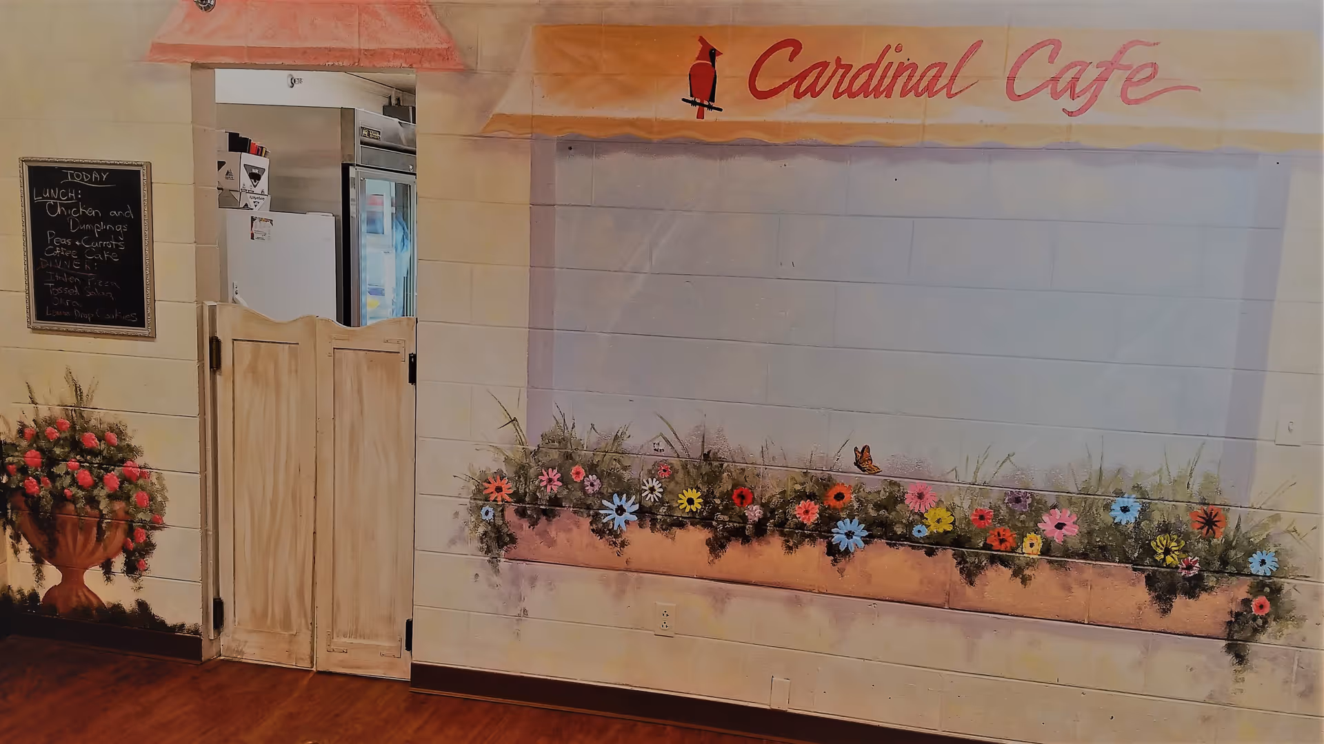 Interior view of Cardinal Cafe with a painted wall mural featuring colorful flowers and a cardinal bird logo. There is a small swinging door leading to a kitchen area with visible refrigerators. A chalkboard menu lists lunch and dinner options.