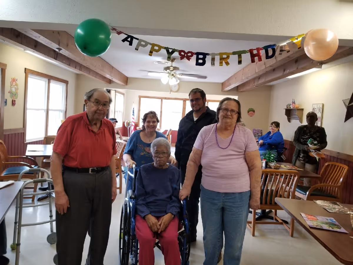 A group of senior residents and staff pose in a decorated common dining room with a 'Happy Birthday' banner and balloons.