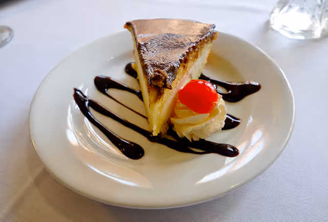 A slice of cheesecake on a white plate, garnished with chocolate syrup drizzles, a dollop of whipped cream, and a red cherry on top, placed on a white tablecloth.