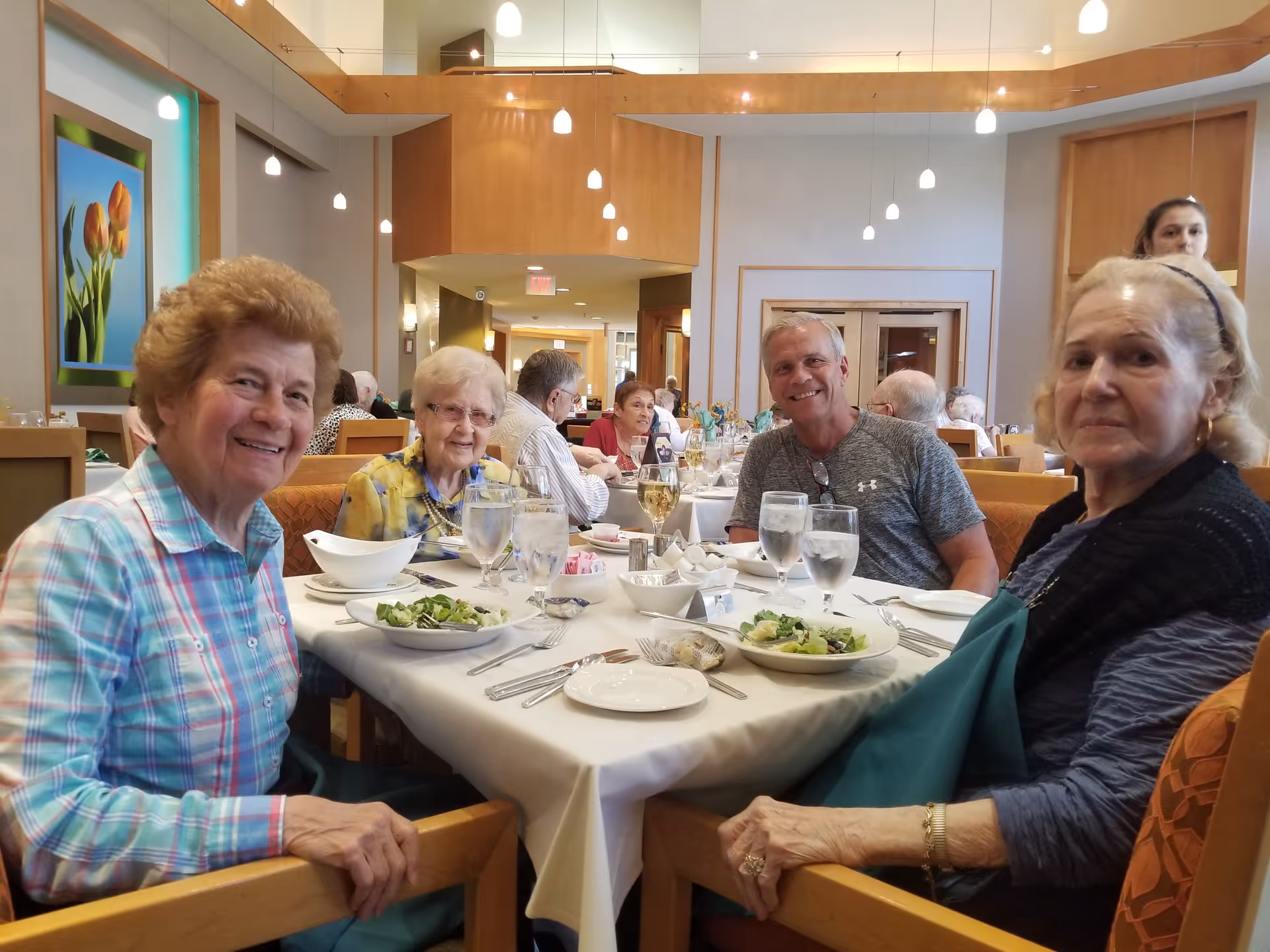 A group of elderly people sitting around a dining table in a well-lit dining room, enjoying a meal with plates of salad and glasses of water and wine. The room has modern pendant lights and wooden accents, with other diners visible in the background.