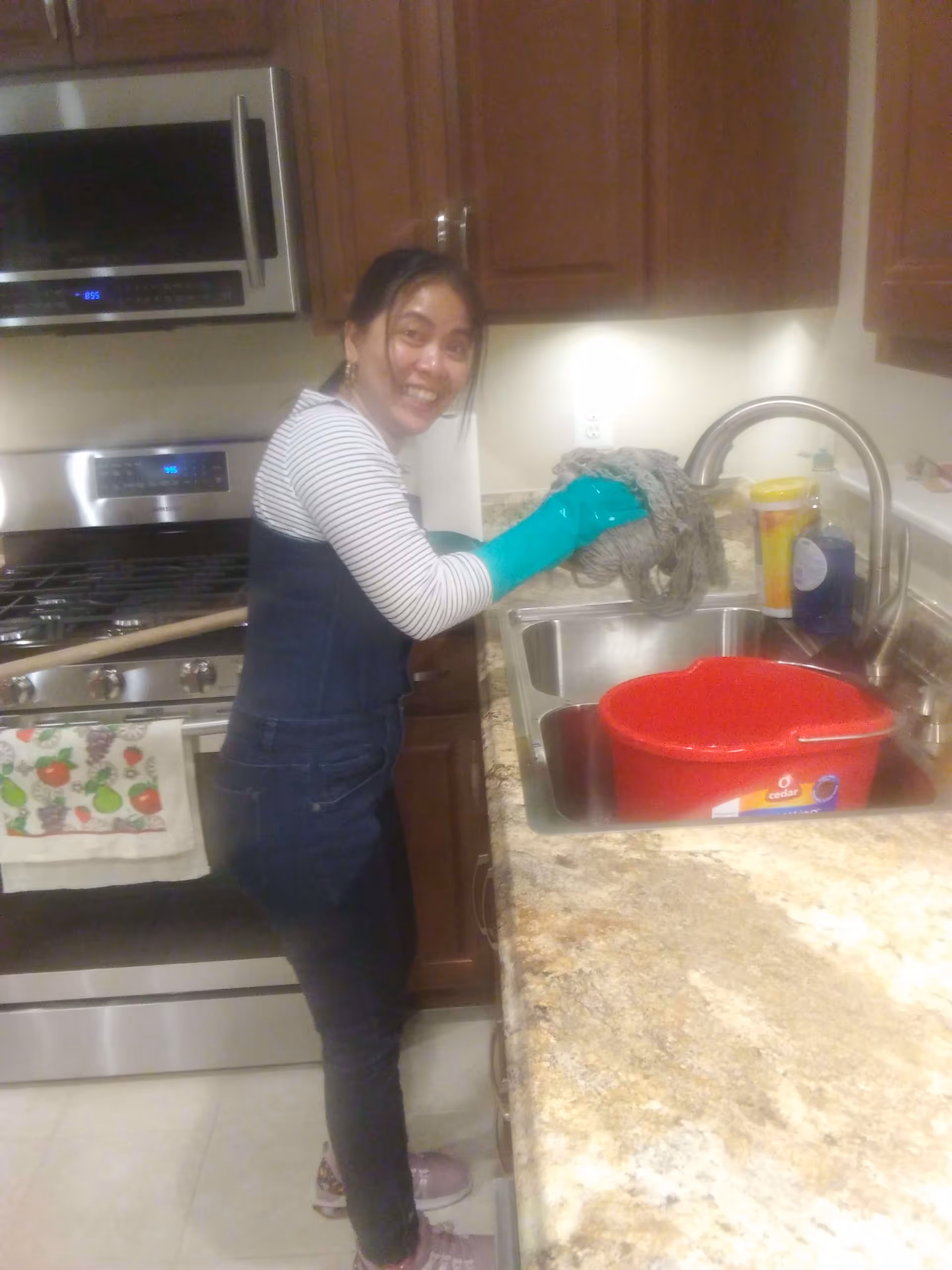 A woman wearing green rubber gloves is standing in a kitchen next to a sink, holding a mop head over the sink. She is smiling and looking at the camera. The kitchen has wooden cabinets, a stainless steel stove and microwave, and a granite countertop with a red bucket in the sink.