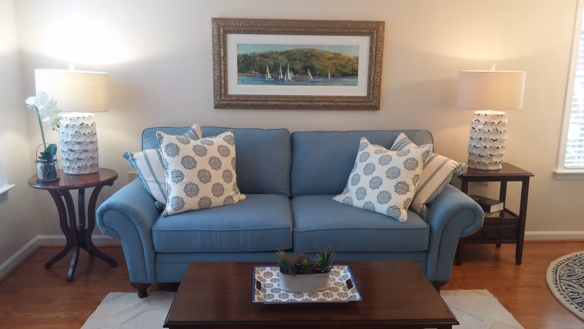 A cozy living room with a blue upholstered sofa adorned with patterned and striped pillows. On either side of the sofa are wooden side tables, each with a white textured lamp and decorative items. Above the sofa hangs a framed painting of sailboats on water with a wooded shoreline in the background. A wooden coffee table in front of the sofa holds a decorative tray with small potted plants. The room has hardwood flooring and light-colored walls with a window on the right side.