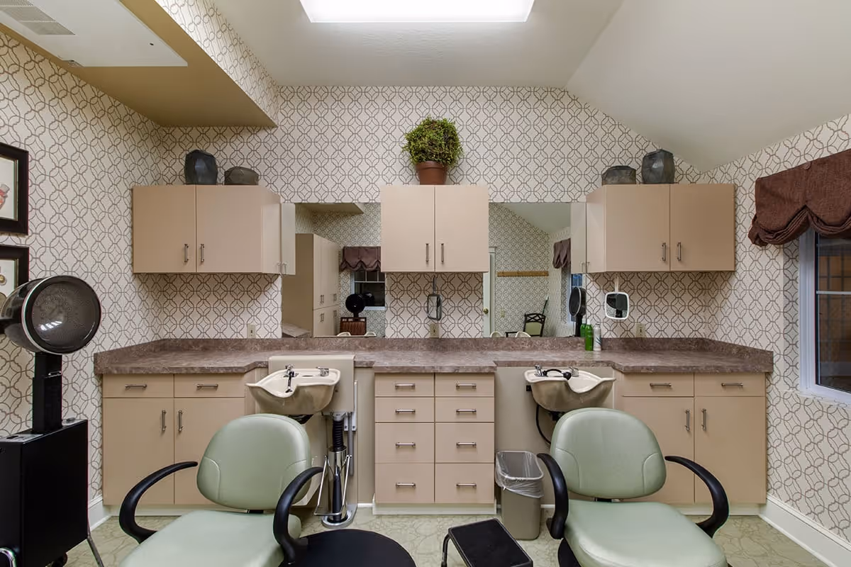 Interior view of a hair salon area in an assisted living facility featuring two green salon chairs with armrests, two hair washing sinks, beige cabinets with drawers and cupboards, a countertop, a wall-mounted mirror, patterned wallpaper, a window with a brown valance, and a hair dryer on a stand.