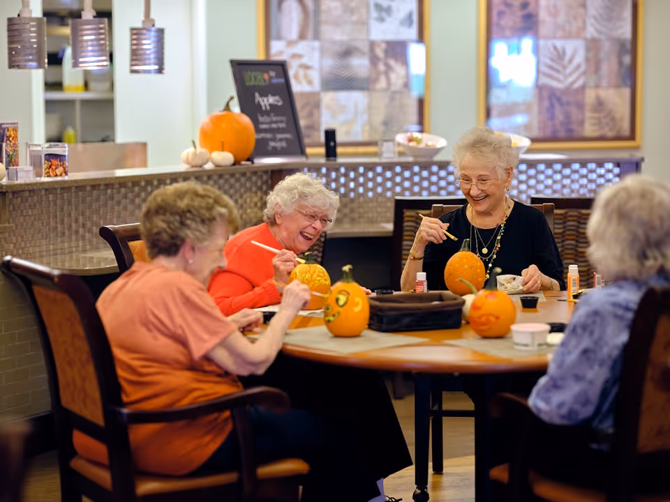 Four elderly women sit around a table in a communal room happily painting and decorating small pumpkins.