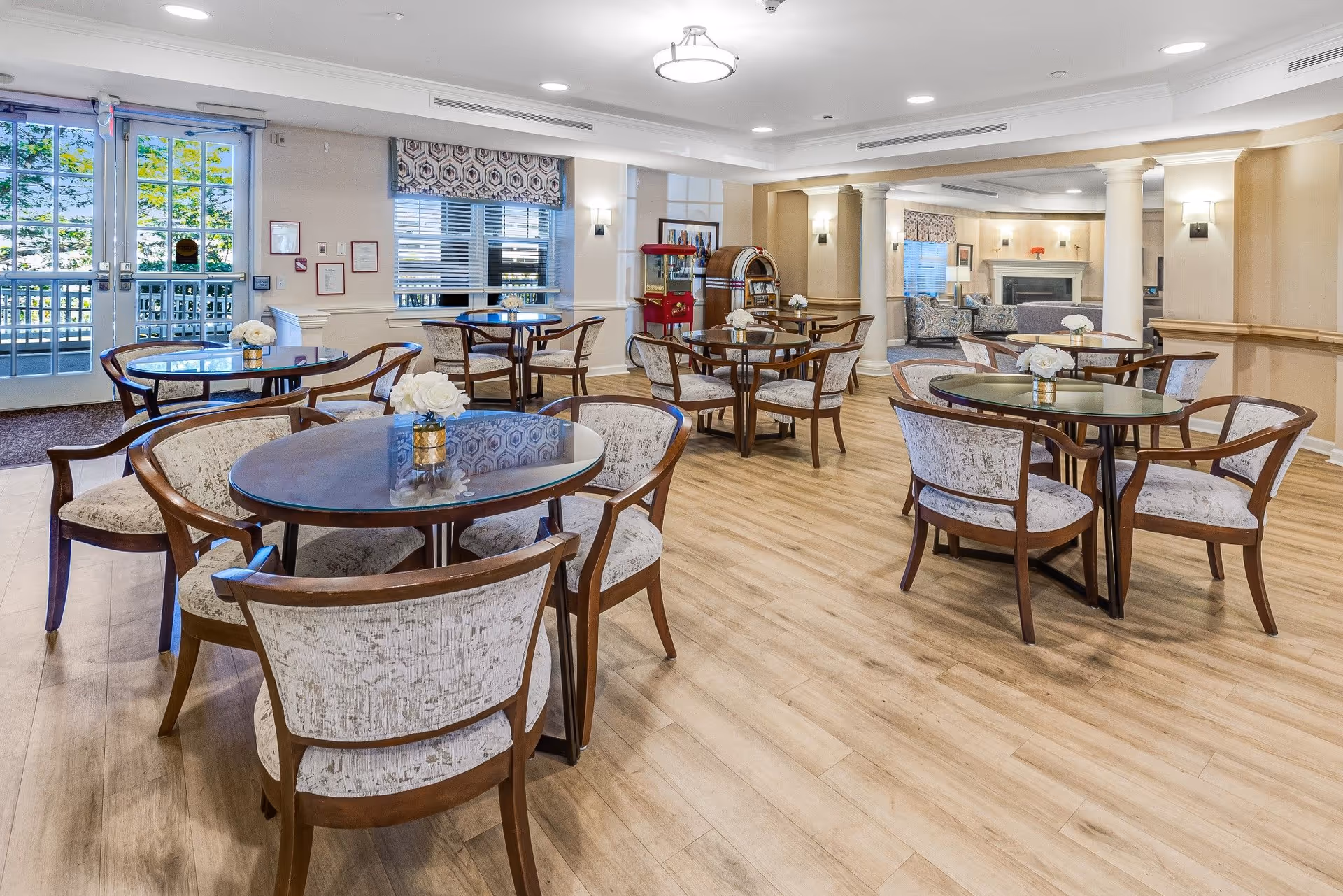 A bright and spacious dining room with several round tables surrounded by cushioned wooden chairs. Each table has a small floral centerpiece. The room features light wood flooring, beige walls, large windows with patterned valances, and a glass door leading outside. In the background, there is a cozy seating area with a fireplace and a vintage jukebox.