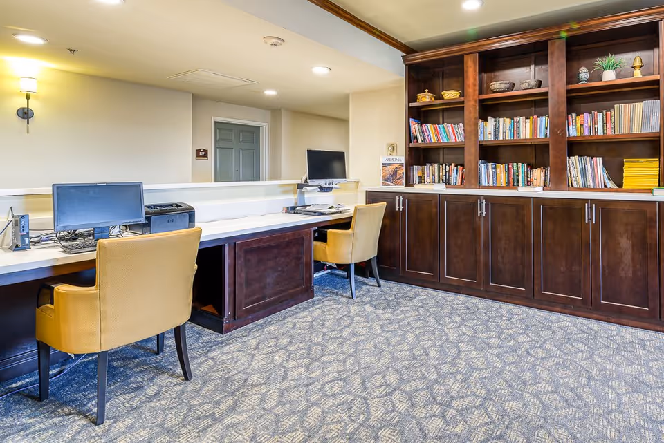 A quiet interior room with two computer workstations, each with a monitor, keyboard, and printer, accompanied by yellow chairs. To the right, there is a large wooden bookshelf filled with books and decorative items. The room has carpeted flooring and soft lighting from a wall sconce and ceiling lights.