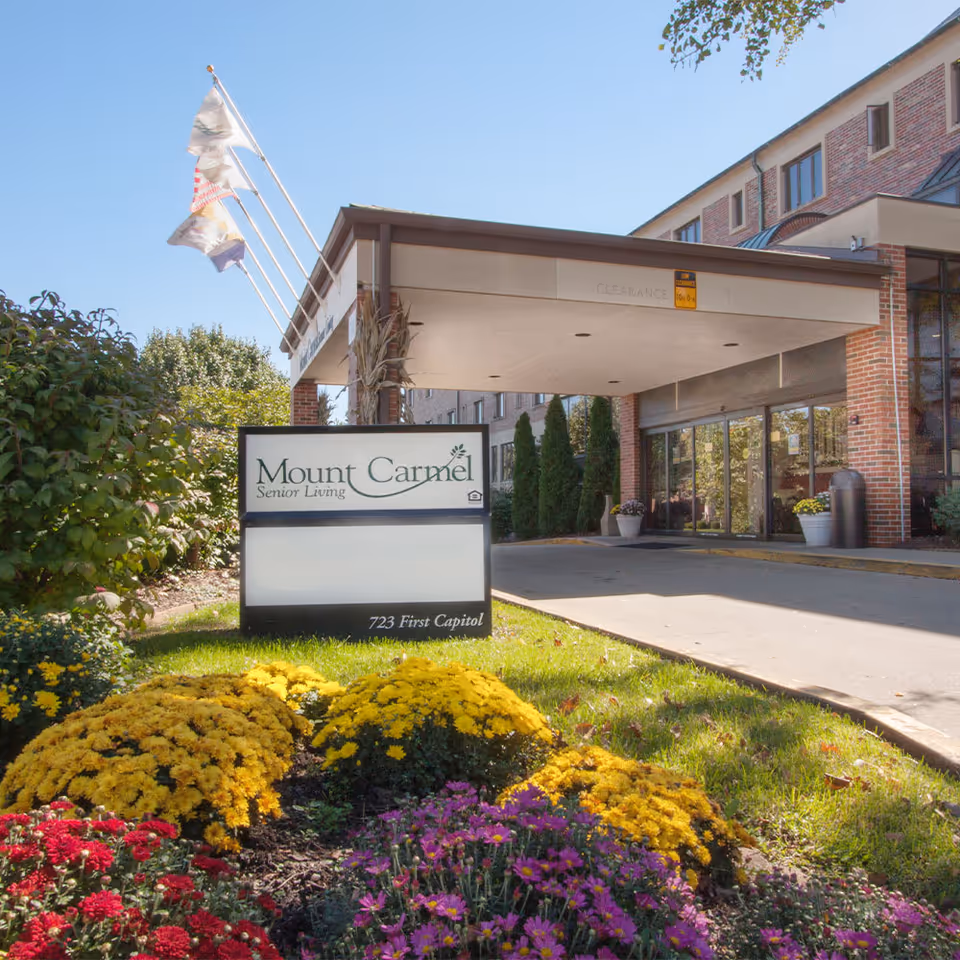 Entrance of Mount Carmel Senior Living facility with a covered drop-off area, a sign displaying the facility name and address, and colorful flower beds in the foreground under a clear blue sky.