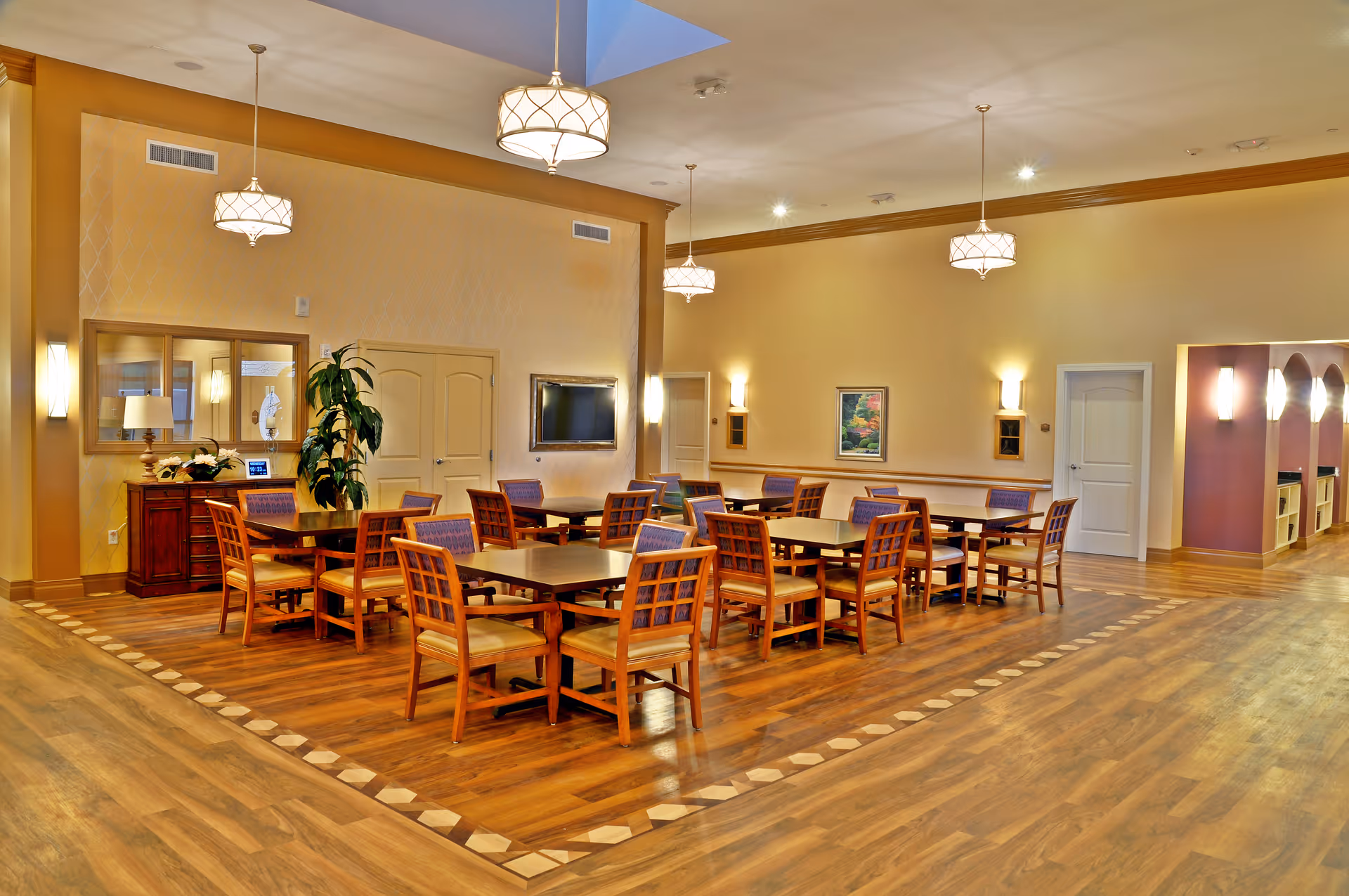 A spacious dining room with multiple wooden tables and chairs arranged neatly on a patterned wooden floor. The room is well-lit with hanging pendant lights and wall sconces. There is a large plant near a sideboard with a lamp and decorative items. A flat-screen TV is mounted on the wall, and a framed picture hangs beside it. The walls are painted in warm tones, and there is a hallway visible to the right with arched openings and additional lighting.