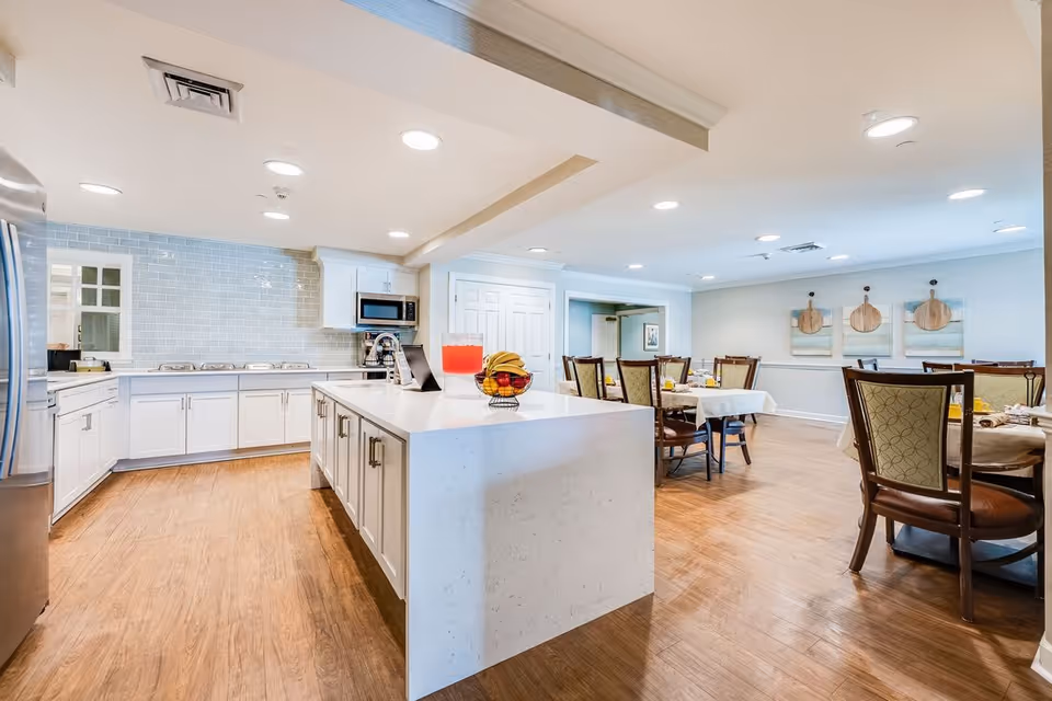 A bright and spacious kitchen and dining area in a senior living facility. The kitchen features white cabinets, a large white island with a bowl of fruit and a pitcher of red beverage, stainless steel appliances, and a light blue tiled backsplash. The dining area has several tables set with tablecloths and chairs, with decorative wall art featuring wooden cutting boards.