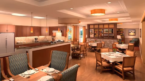 A spacious dining area with several wooden tables and chairs, some tables set with white plates and cups. The room features warm lighting with ceiling fixtures and pendant lights over a kitchen island. The kitchen area includes wooden cabinets, a refrigerator, and a coffee maker. In the background, there is a cozy seating area with bookshelves and framed artwork on the walls.
