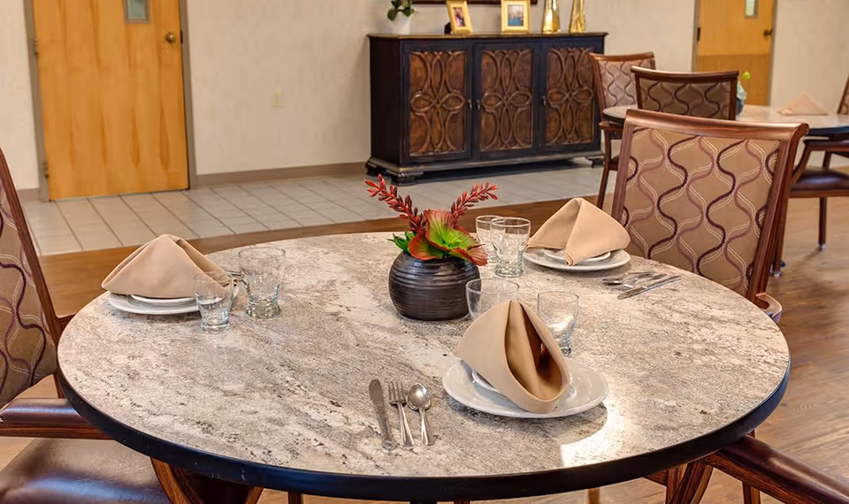 Round dining table set with plates, folded napkins, glasses and a floral centerpiece in a communal dining room.