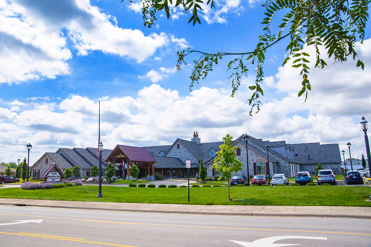 Front view of The Inn at Winchester Trail, a stone senior living building with pitched roofs, a parking lot, and landscaped lawn under a blue sky.