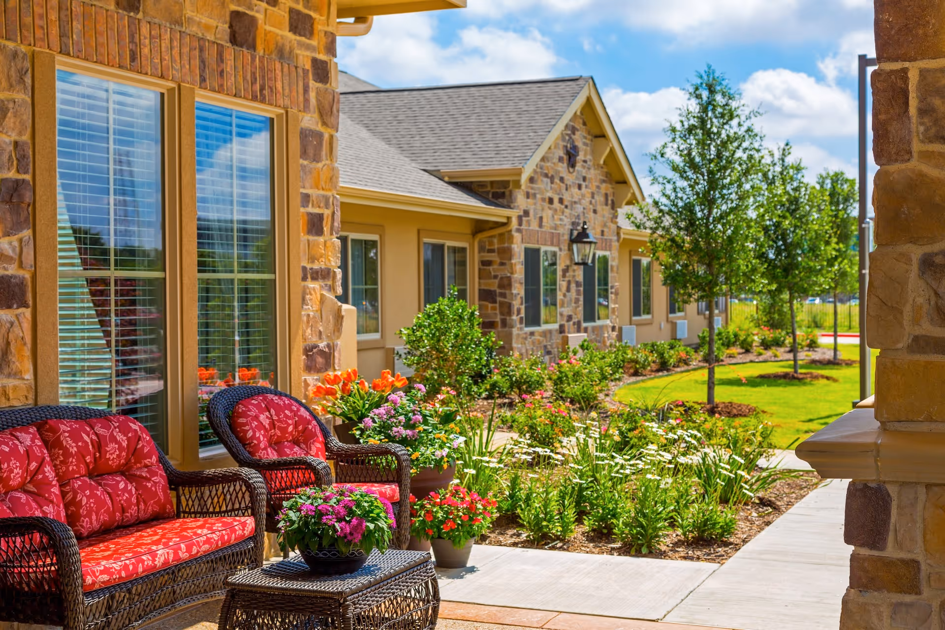 Outdoor patio area at Arbor Hills Memory Care Community featuring wicker furniture with red floral cushions, a small wicker table with a potted plant, and a landscaped garden with various flowers and trees along a concrete walkway beside a stone and stucco building.