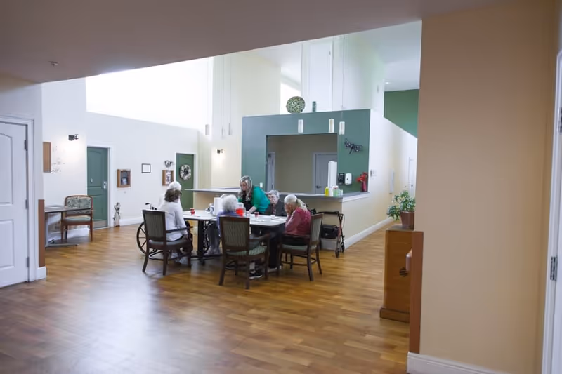 A group of elderly people sitting around a table in a spacious, well-lit common area with wooden flooring and light-colored walls. A caregiver is interacting with them. The room has green doors and some wall decorations, with a counter area in the background.