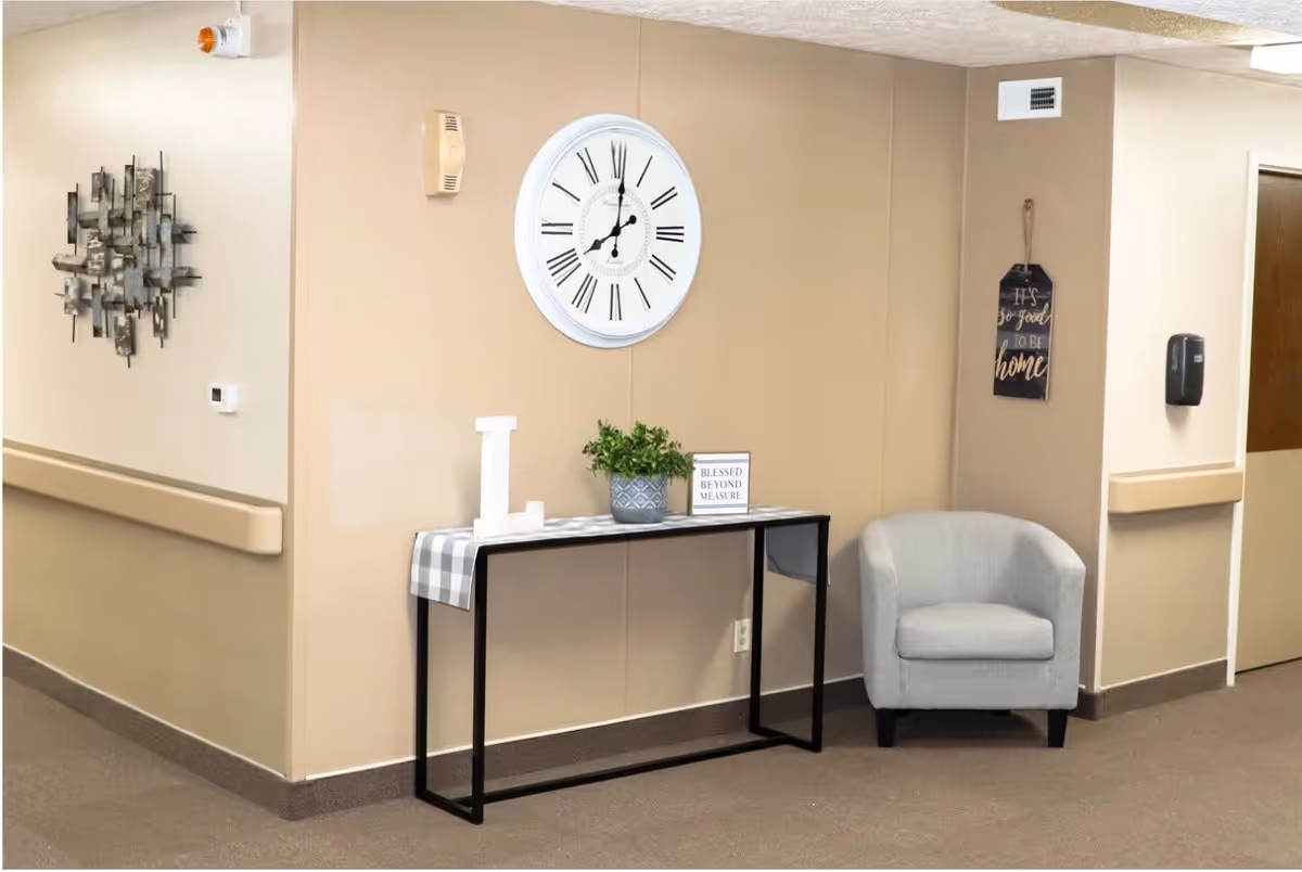 A hallway corner in a senior living facility with beige walls and carpeted floor. A small black metal table with a checkered runner holds a white letter L, a potted plant, and a sign that reads 'BLESSED BEYOND MEASURE.' Above the table is a large white wall clock with Roman numerals. To the right of the table is a light gray upholstered armchair. Wall decor includes a metal abstract sculpture on the left wall and a hanging sign on the right wall that says 'IT'S so good TO BE home.'