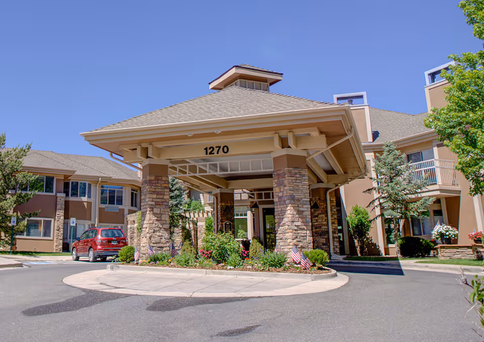 Entrance of a senior living facility with a covered driveway supported by stone pillars, surrounded by landscaped greenery and small American flags, with a clear blue sky overhead.