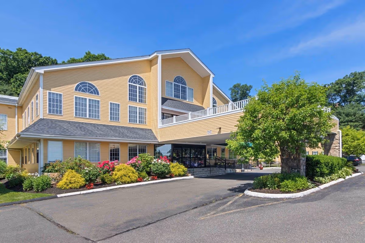 Exterior view of a two-story yellow building with large windows and a covered entrance. The building is surrounded by landscaped bushes, flowers, and trees under a clear blue sky.