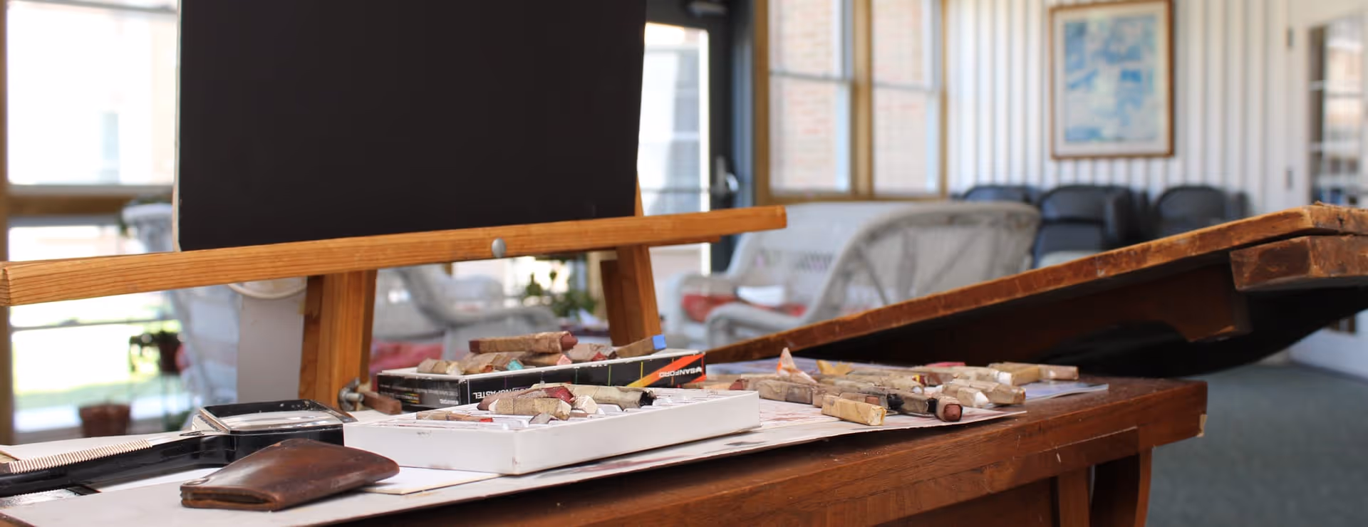 Close-up of an artist's workspace with pastel sticks and a blank black canvas on an easel in a bright room with wicker chairs and large windows in the background.