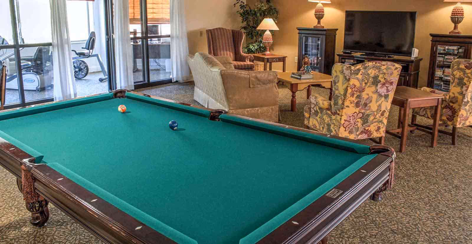 A recreational room with a green felt pool table in the foreground, two billiard balls on the table, and a seating area in the background featuring a sofa, floral armchairs, a coffee table, a TV on a stand, and two lamps on side tables. Large windows with sheer curtains reveal exercise equipment outside.
