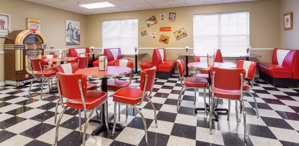 A retro-style dining room with red and white chairs and booths, black and white checkered floor, vintage posters on the walls, and a classic jukebox in the corner.