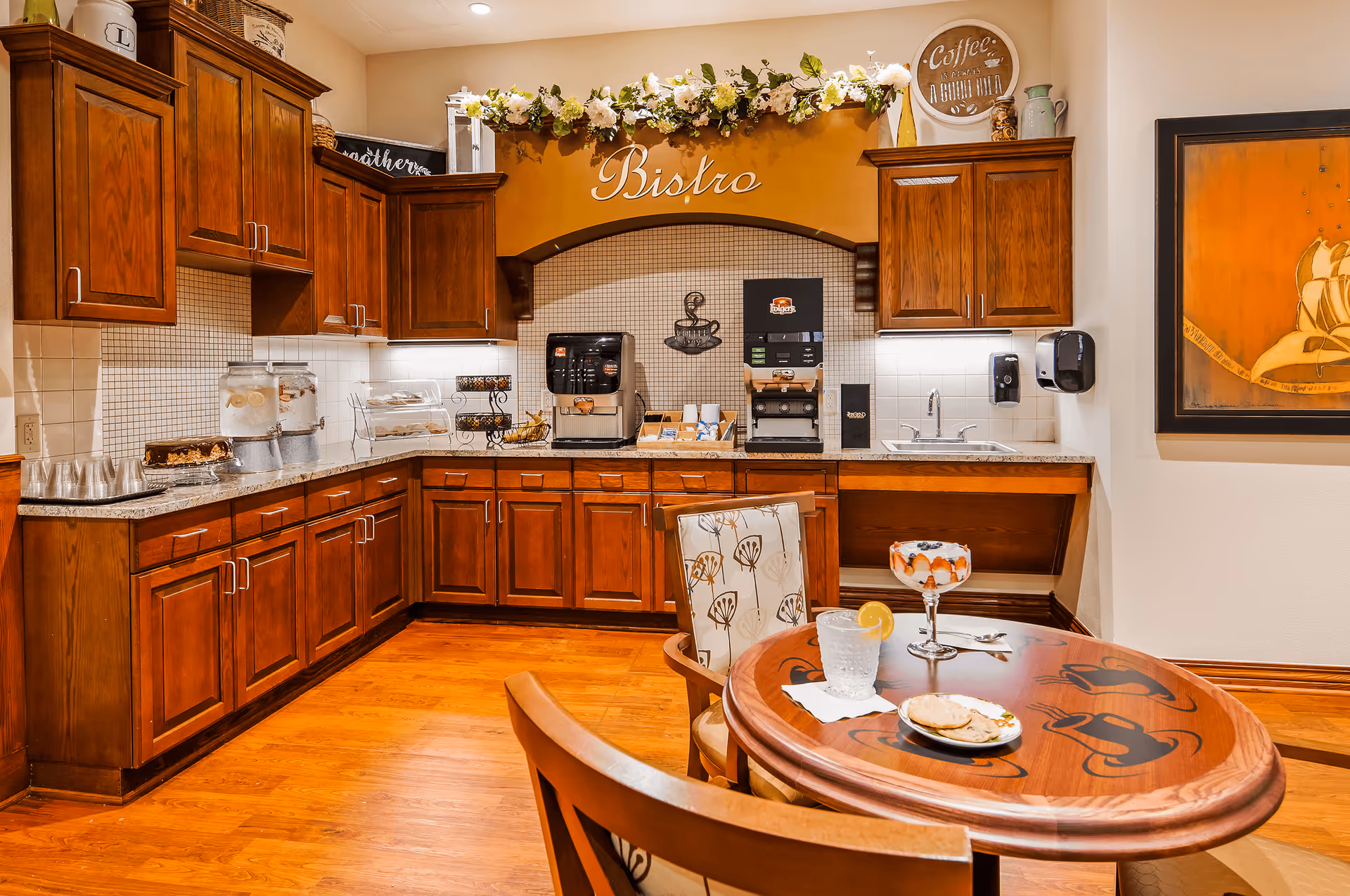A cozy bistro area with wooden cabinets and a countertop featuring a coffee machine, water dispensers, and snacks. A round wooden table with coffee cup designs is set with a glass of water with lemon, a dessert parfait, and a plate of crackers and cheese. The walls are decorated with floral garlands and framed artwork.
