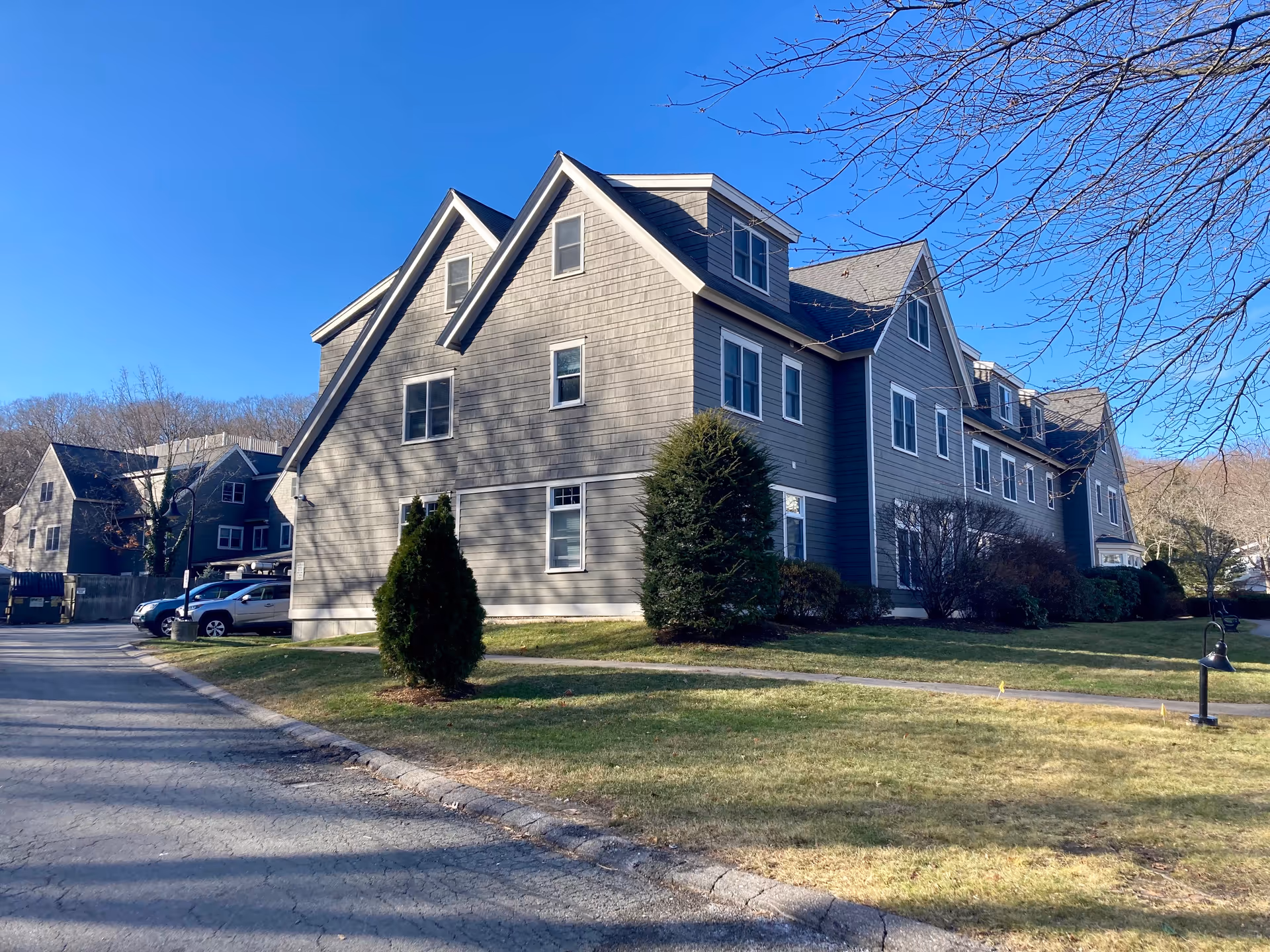 Three-story gray shingle senior living building with dormer windows, a lawn, and parked cars under a clear blue sky.
