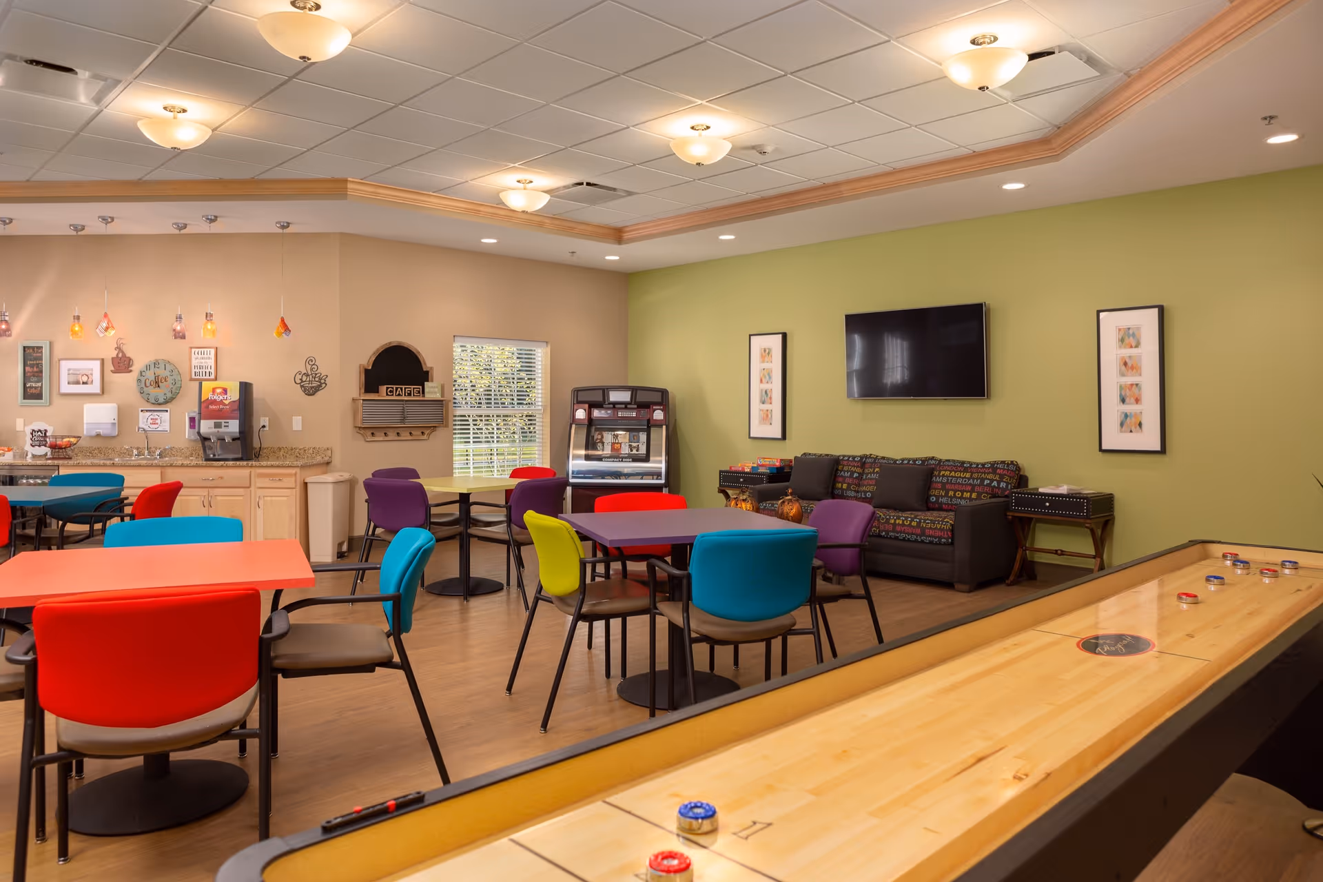 Colorful senior living common room with tables and chairs, a shuffleboard table, kitchenette area, couch and wall-mounted TV.