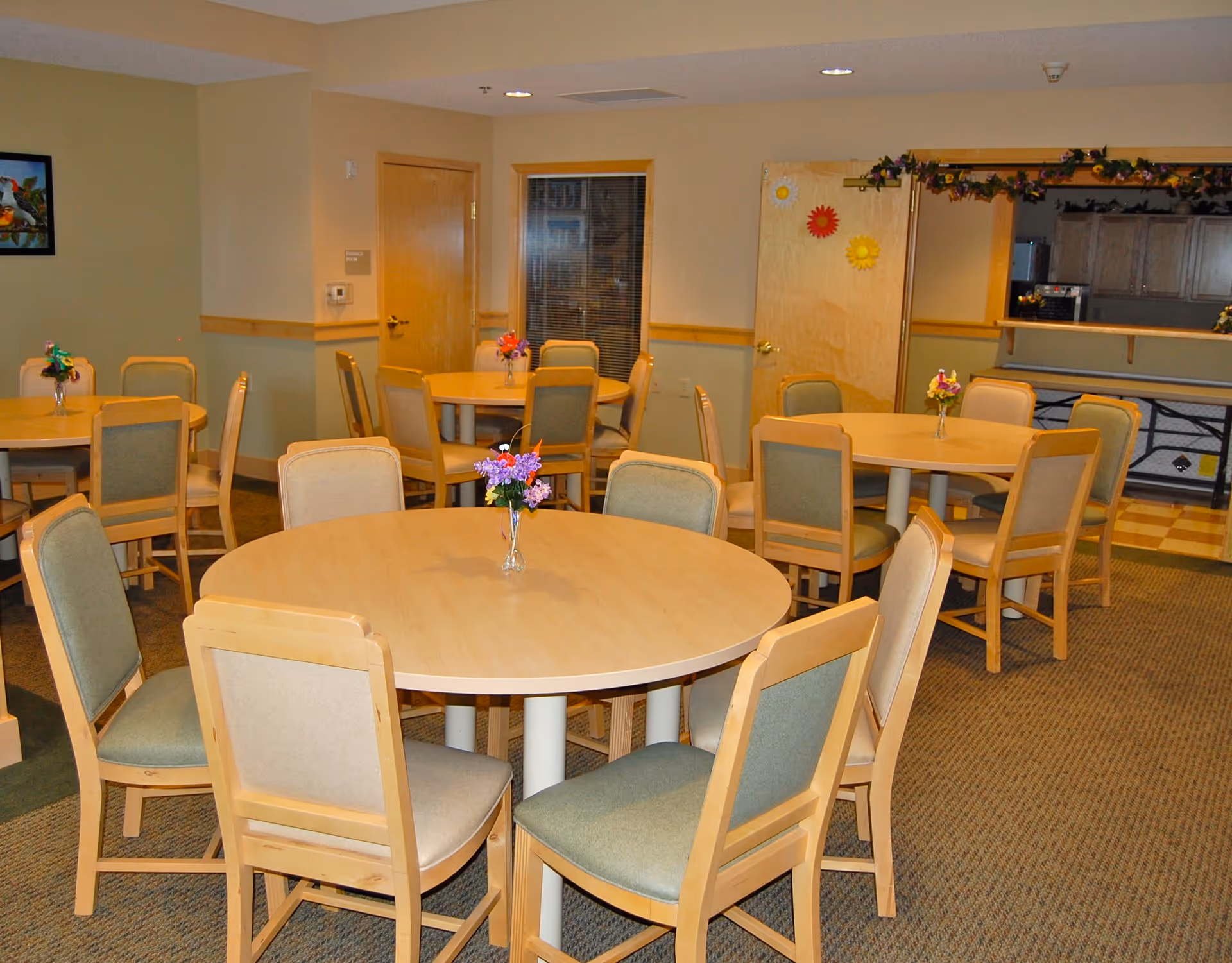 A senior living facility dining room with multiple round wooden tables surrounded by cushioned chairs. Each table has a small vase with colorful flowers. The room has beige walls, a carpeted floor, and a serving window to a kitchen area in the background. There are doors and a window visible on the far wall, and some decorative flowers hanging above the serving window.