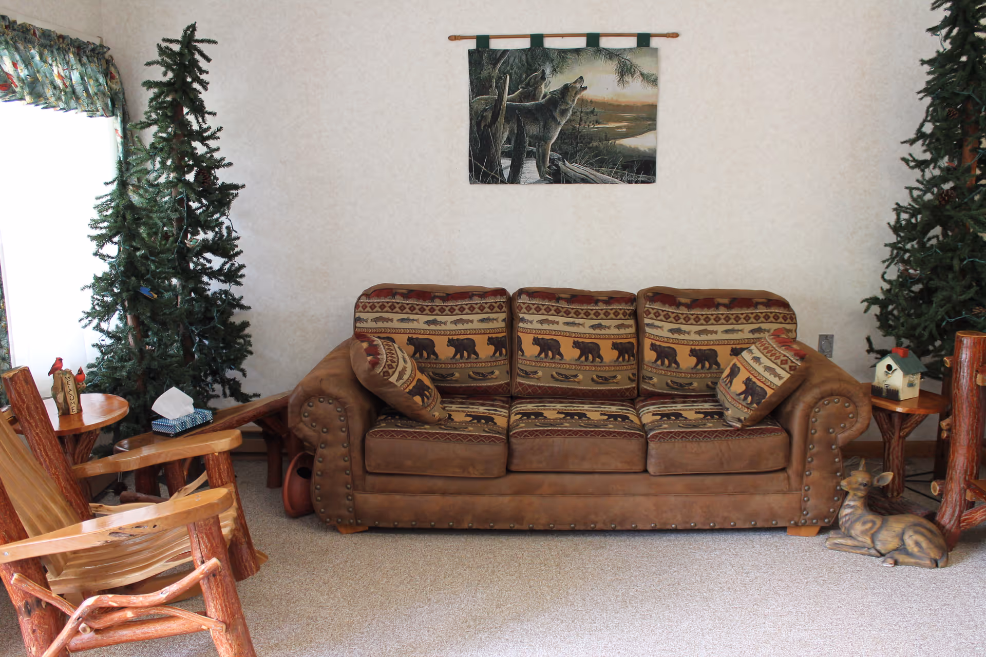 A cozy living room with a brown leather sofa featuring bear-patterned cushions, flanked by two artificial pine trees. There are rustic wooden chairs and side tables on either side of the sofa. A wall hanging depicting wolves in a natural setting is centered above the sofa. A small decorative deer figurine is placed on the floor to the right.