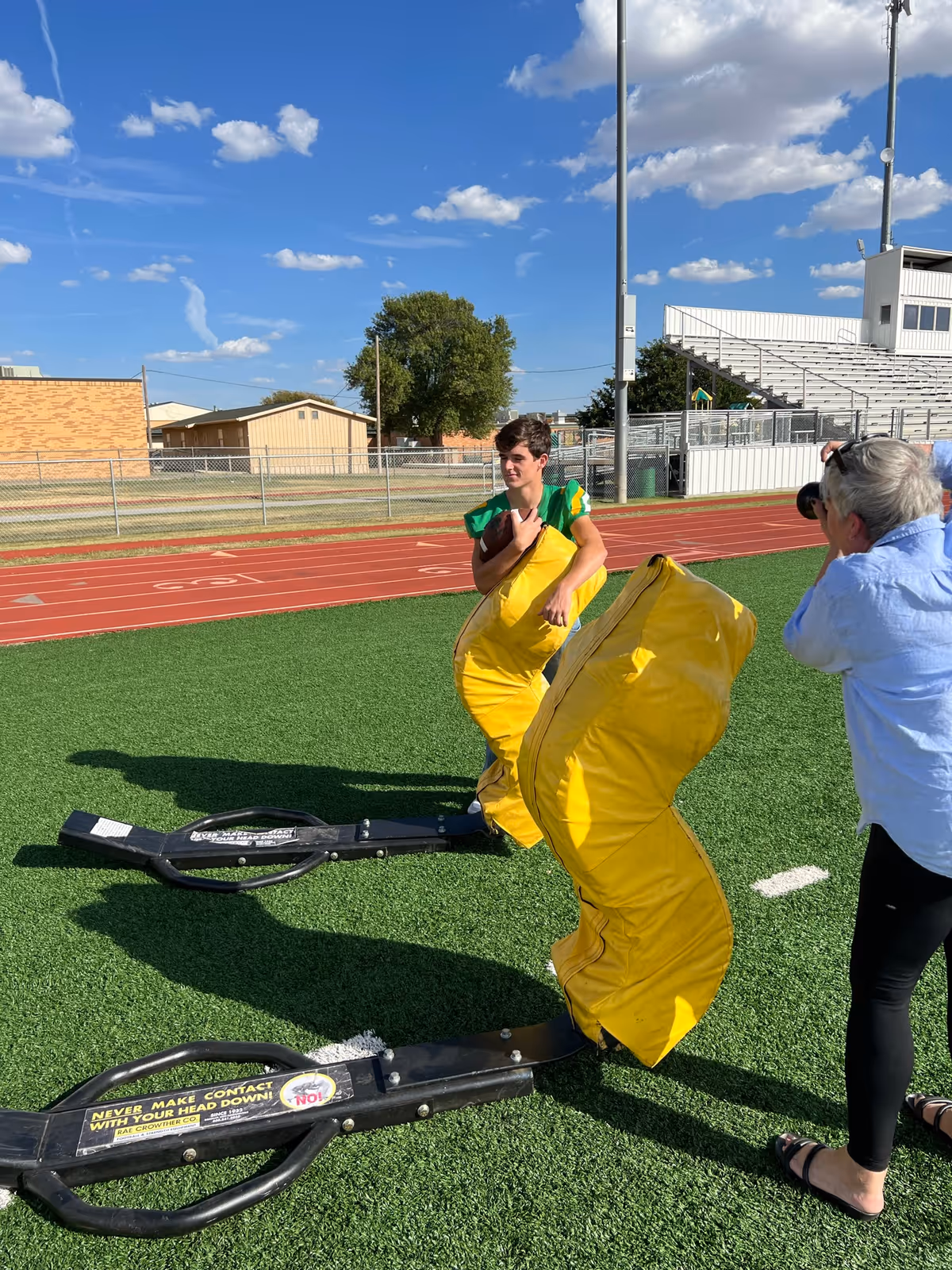 A young man in a green and yellow sports uniform stands on a football field holding a large yellow padded blocking sled. A woman with gray hair, wearing a light blue shirt and black pants, is taking a photo of him. The scene is outdoors on a sunny day with a blue sky and scattered clouds. In the background, there is a running track, bleachers, and some buildings.