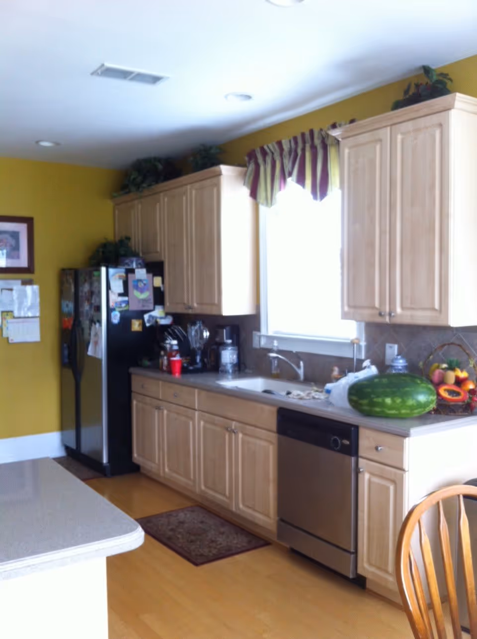 A kitchen with light wood cabinets, a stainless steel dishwasher, and a black refrigerator covered with magnets and papers. There is a large watermelon and a basket of fruit on the countertop next to the sink. A window with a striped valance is above the sink, and a wooden chair is partially visible in the foreground.