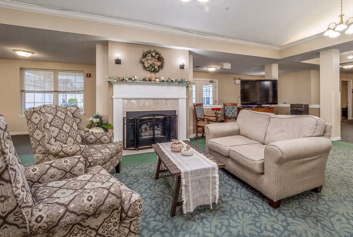 A cozy living room area in The Addison of Moorehead Place featuring a beige couch, two patterned armchairs, a wooden coffee table with decorative items, a fireplace with a wreath and garland above it, and a TV on a stand in the background.