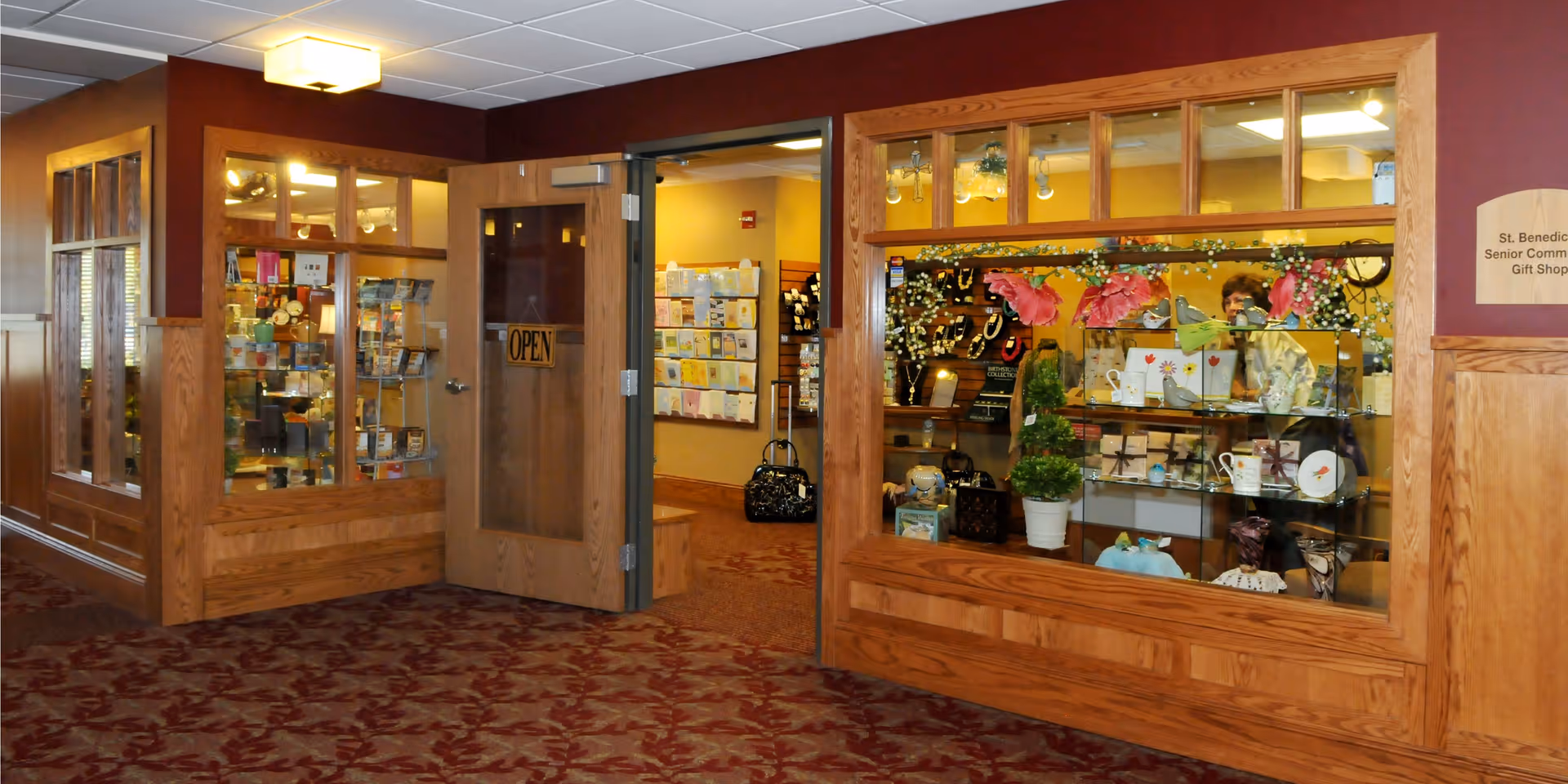 Interior view of a senior living facility gift shop with wooden framed glass windows and an open door. Inside the shop, various gift items such as bags, mugs, and decorative pieces are displayed on shelves. The floor is carpeted with a red and brown patterned design, and the walls are painted in warm tones.