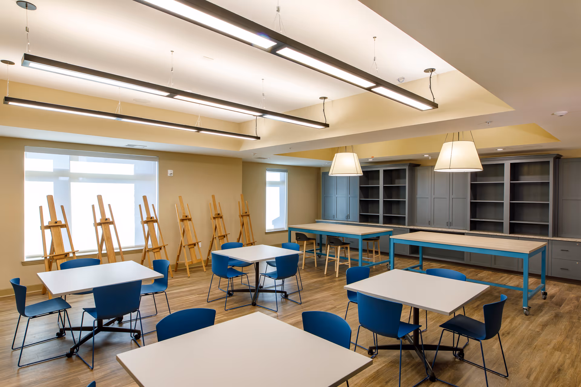 A spacious, well-lit room with several square tables surrounded by blue chairs. Along one wall, there are multiple wooden easels lined up in front of large windows. Two long tables with blue legs and wooden tops are positioned near built-in gray cabinets with open shelving. The ceiling features modern hanging light fixtures.