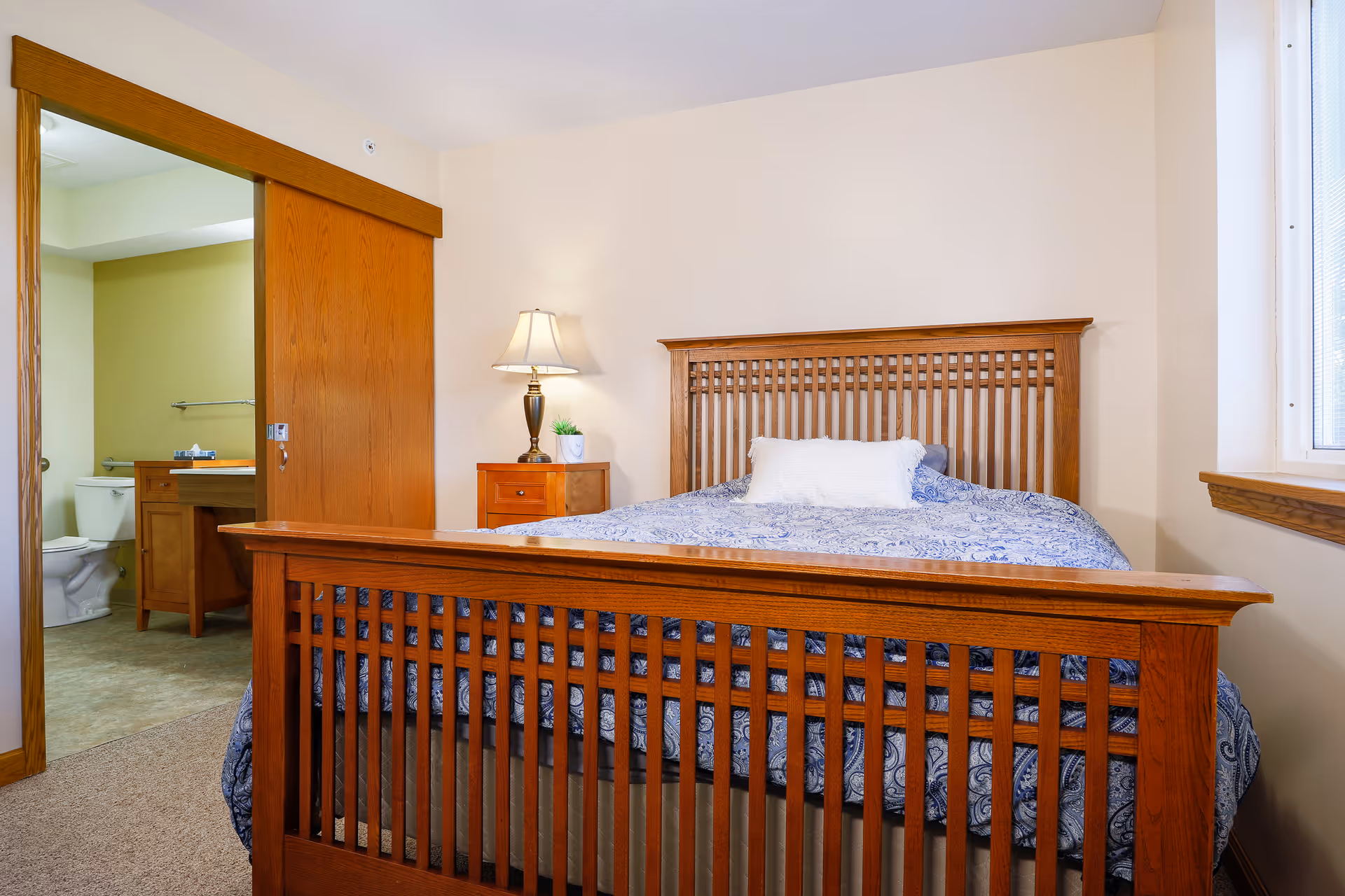 Wooden bed with blue patterned bedding and a nightstand lamp in a bedroom with a sliding door open to an en-suite bathroom.