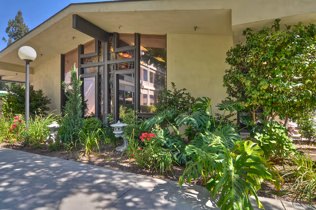 Exterior view of a building corner at California Mission Inn - Rose Manor with large windows, beige walls, and a garden area featuring various green plants and red flowers. A round white lamp post is visible on the left side along a paved walkway.