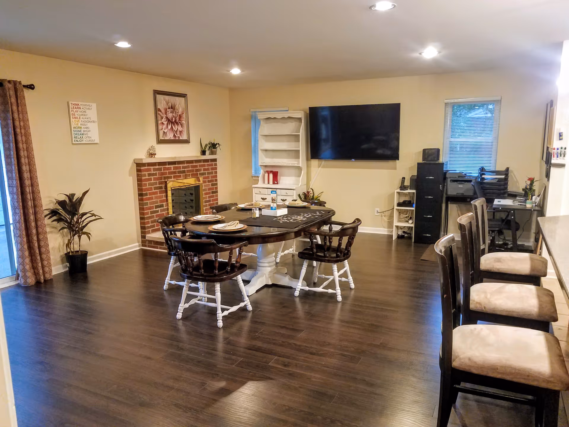 A cozy living room area with a dark wooden dining table set with plates and utensils, surrounded by four chairs with white legs and dark wooden seats and backs. The room features a brick fireplace with a floral painting above it, a white cabinet, a large flat-screen TV mounted on the wall, and two windows with blinds. There is a small office area with a filing cabinet, printer, and desk with a chair on the right side. The floor is dark wood, and the walls are painted beige. Three bar stools with cushioned seats are lined up along a counter on the right side.