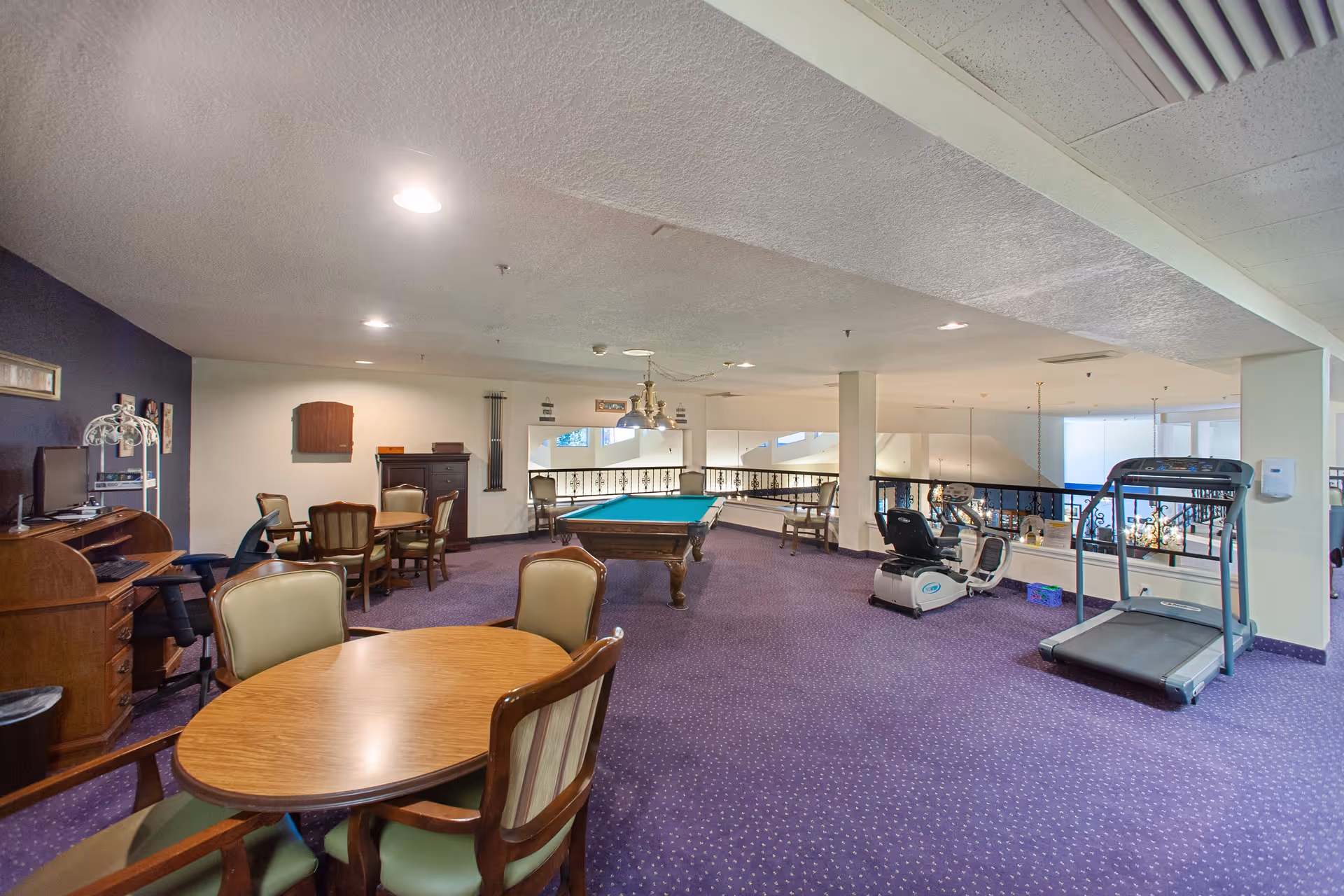 A spacious common area with purple carpet featuring a pool table in the center, a treadmill and exercise bike on the right, and several wooden tables and chairs on the left. The room has a low ceiling with recessed lighting and a railing overlooking a lower level.