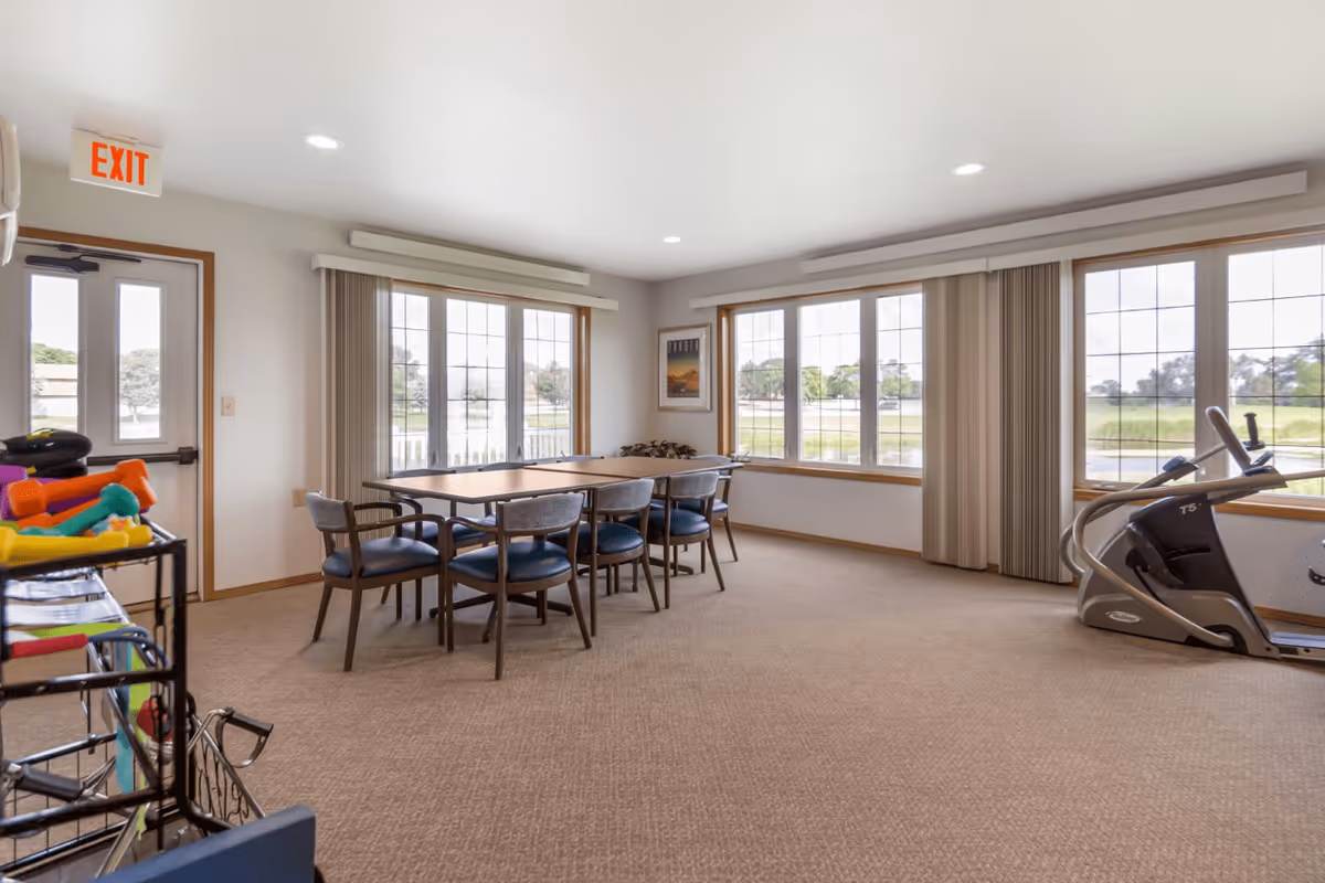 A bright room with large windows letting in natural light, featuring a rectangular table with six chairs around it. To the right, there is exercise equipment including a treadmill. On the left side, there is a rack with colorful dumbbells and other exercise accessories. An exit door is visible on the left wall.