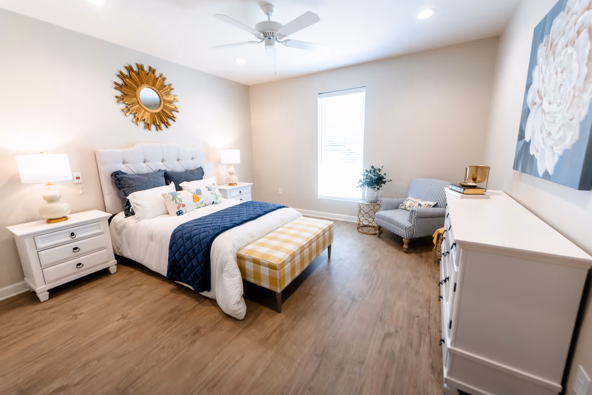 A bright and neatly arranged bedroom with a large bed featuring white and navy bedding, two white nightstands with lamps, a yellow checkered bench at the foot of the bed, a striped armchair next to a window, a white dresser with books and a decorative item, a ceiling fan, and a sunburst mirror on the wall above the bed.