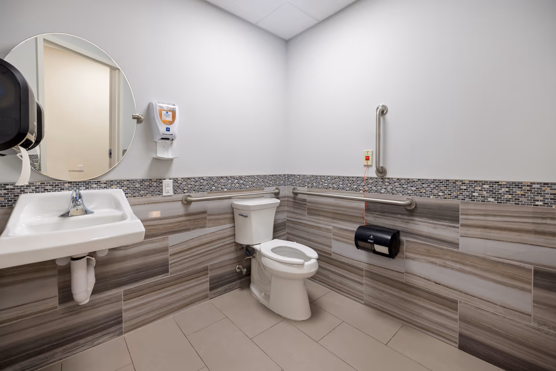 A clean and accessible bathroom with a white toilet, a wall-mounted sink, a round mirror, handrails on the walls, a soap dispenser, and a toilet paper holder. The walls have a decorative tile border and the floor is tiled.