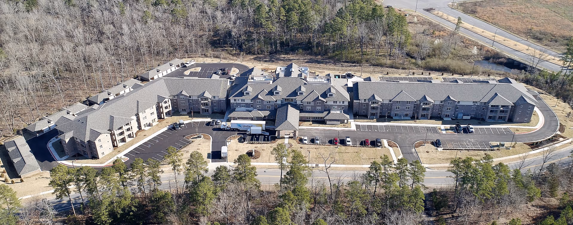 Aerial view of a large, multi-wing retirement facility surrounded by trees and parking lots. The building has multiple entrances and a covered drop-off area at the front. The surrounding area includes roads and wooded land.